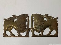 Two brass lion wall decor pieces shown together facing each other on a white background, detailed stamped brass with swords and artistic patterns visible.