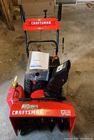 Front view of snow thrower with gas can and owner's manual