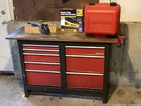 Red and black Craftsman metal tool chest workbench with multiple drawers against concrete wall