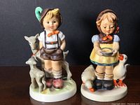 Front view of two Goebel Hummel figurines: a boy with goats and a girl feeding ducks, both standing on bases.