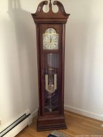 Full view of the wooden grandfather clock standing on a wooden floor near a heater baseboard. Shows overall size and design with glass door, pendulum, and weights visible inside.