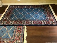 Full view of one blue area rug laid flat on wood floor showing central lattice and floral pattern with ornate border and cream fringe edges.