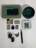Top-down view displaying 2 ceramic ash receivers (rectangular and round), one white ceramic ashtray labeled 'Cairo Shepheards Hotel', 5 metal lighters in varied colors, matchbox, and 2 wooden pipes.