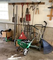 Yard tools displayed against a garage wall including shovels, rakes, a green broadcast spreader, and an assortment of handles and tools inside boxes.