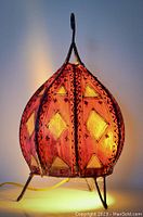 Arabesque style leather-covered table desk lamp lit, showing glowing red leather with diamond yellowish cutouts and metal framing, on three metal legs.