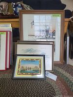 Five framed artworks arranged with two standing and three leaning on carpet, showing visible wrinkles and scuffed frames