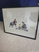 Side perspective of framed Asian artwork showing three figures in traditional attire, set against a light background, in black metal frame with paint wear.