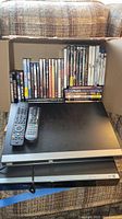 Full lot contents in cardboard box on couch, showing media players, remote controls, and DVDs