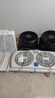 Four small fans arranged on carpet: Haier box fan, Holmes dual fan window unit, Honeywell box fan, and two black cyclone fans.