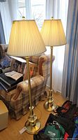 Three brass floor lamps with pleated white fabric lampshades, placed next to a couch. Two lamps are matching, one differs slightly in base shape.