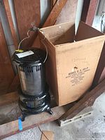 Metal kerosene stove/heater standing next to its original cardboard box with visible labels and handwritten notes.