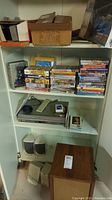 Stack of DVDs and VHS tapes with Zenith DVD/VHS combo player and speakers below on white shelf