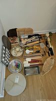 Overview of baking pans, utensils, wooden spoons, spatulas, measuring cups, wire basket, baking trays, and bag clips arranged on floor.