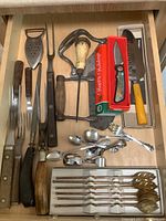 Drawer view of assorted vintage kitchen utensils including cake server, meat forks, pocket cutter in box, peeler, and various spoons and forks