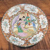 Top view of porcelain plate showing traditional Asian artistic design with two women, floral motifs, and gold trim.