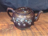 Front view of antique brown stoneware teapot showing floral design, handle, spout, and lid on top