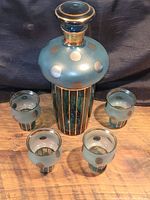 Full view of teal blue crystal decanter with gold pin stripes and matching five glasses with gold polka dots, displayed on wooden surface against dark background.