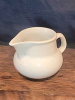Front side view of an off white glazed pottery cream or milk pitcher on a wooden surface