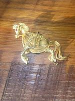 Gold tone horse brooch pin resting on a wooden surface, showing three-dimensional wire and cast design with ruby stone eye and pin backing.