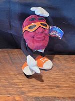 Full view of the plush California Raisin collectible toy, showcasing the front with orange sunglasses, white gloves and shoes, and attached tags.