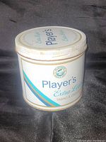 Photo of a white cylindrical metal 'Player's Extra Light' tobacco tin with blue and gold stripes and a screw-on lid, standing upright against a black background.