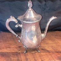 Vintage solid brass silver plated coffee pot with ornate handle, curved spout, and decorative finial lid, standing on four feet, photographed on wooden surface with dark cloth background.