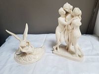 Two ivory-colored classical sculpture figurines placed together on white cloth background, with gray wall backdrop.