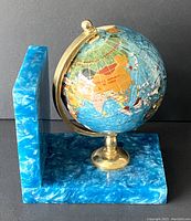 Front and side angled view of the aqua blue pearlized Lucite bookend with a revolving globe attached. The globe shows Asian and African continents with semiprecious stones mounted inlaid into the surface.