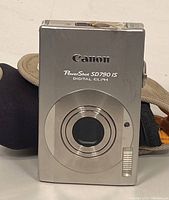 Front view of Canon PowerShot SD790 IS camera showing silver metallic body, lens area, and part of attached case.