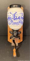 Front view showing ceramic container with blue windmill scene, grinding mechanism, and wooden handle.