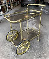 Vintage Hollywood Regency style brass tone bar cart with smoke glass shelves on concrete floor. Shows front corner and side with brass tone spoked wheels and caster wheels.