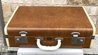 Front view of closed brown vintage suitcase with cream edging and white handle, showing metal latch locks.