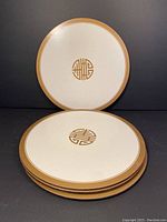 Four round Pimpernel placemats stacked showing front design of gold Shou symbol on cream background with gold rim.