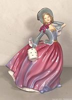 Front and slightly angled view of the Royal Doulton Autumn Breeze figurine showing detailed facial features, the pink and purple flowing dress, blue-grey bodice, hat with pink ribbon, and the white muff.