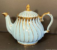 Front view of turquoise ceramic Sadler teapot with prominent gold swirled accents, spout on left, handle on right, gold details on spout tip and handle edges.