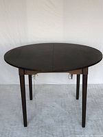 Front view of the round dining table with the folding extension leaf closed, showing the dark wood veneered surface and tapered legs.