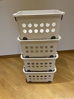 Four stacked white plastic storage baskets with circular holes and wheels on a wooden floor against a plain wall.
