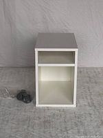 Front view of white IKEA nightstand showing open top compartment and closed bottom compartment with detached wheels beside it.