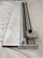 Top view of large format rotary paper trimmer showing white surface with discoloration and metal cutting edge with rotary blade and black knob.