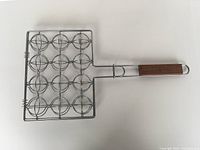 Angle view of closed rectangular stainless steel grilling basket with 12 circular compartments and wooden handle branded 'Sur La Table'.