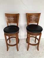 Front view of two wooden swivel bar stools with black leather seats and woven wood backrests side by side.