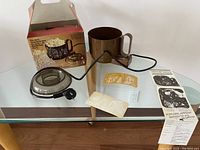 Full display of popcorn maker components including heating base with cord, kettle, plastic lid, original box, and instruction leaflet on glass table.