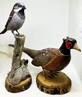 Two wooden bird sculptures displayed against white background, one perched songbird on a wood branch base and one ring-necked pheasant on a bark-textured wooden base. Signed by artist J.D. Rempel.