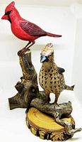 Photo of both wooden bird sculptures together, cardinal on left on tall wood branch base, grouse on right on smaller curved log base
