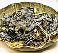 Close-up photo of assorted rhinestone brooches, pins, and some necklaces in a shallow dish showing intricate designs and mixed metal tones.