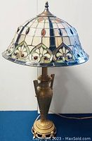 Side-front view of lamp showing stained glass shade with floral and geometric design and metal base with handles and decorative feet.