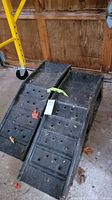 Pair of black metal auto ramps angled upward with perforated surface for traction. Positioned on concrete ground inside a garage with wood walls.
