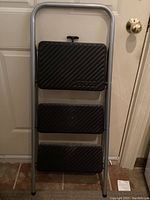 Front view of folded Cosco 3-step ladder showing three black plastic steps and silver metal frame.