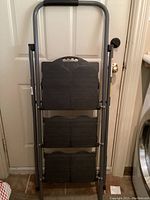 Front view of closed folding Cosco 3-step ladder leaning against door, showing padded handle and ribbed steps in folded position.