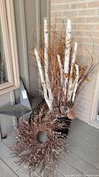 Overall view of the wood branch arrangement with birch branches, twigs, pinecones in black planter and the decorative twig wreath on floor next to it.